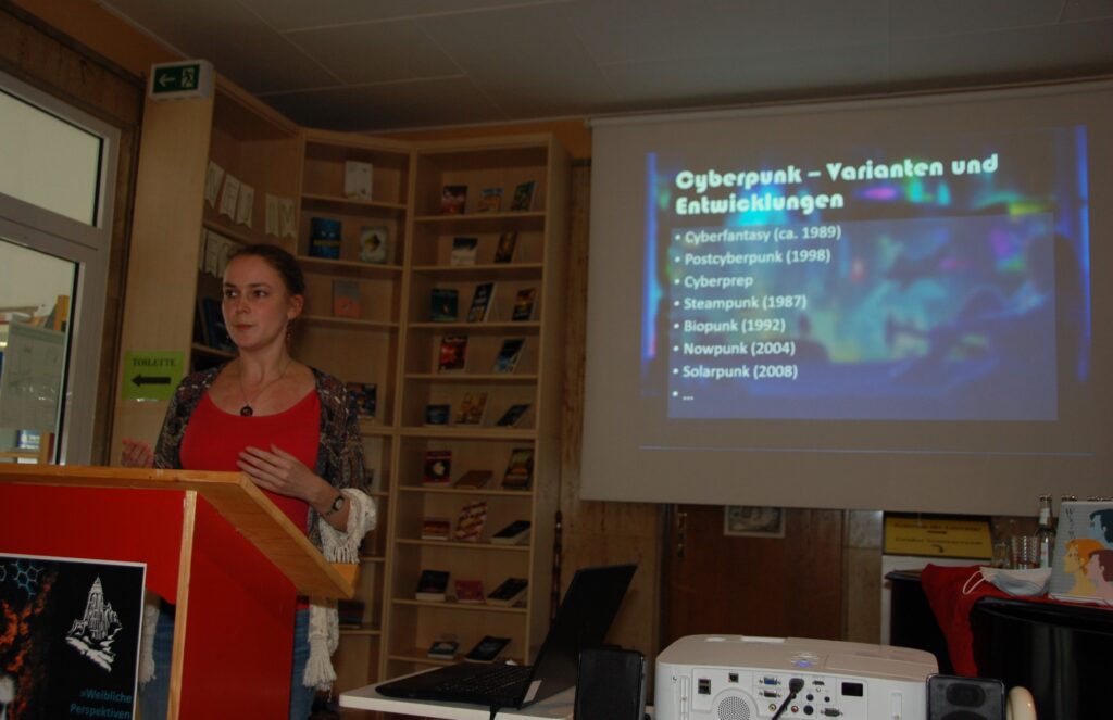 Alessandra Reß vor einer Slide zu Cyberpunk-Varianten und -Entwicklungen. Genannt sind Cyberfantasy (ca. 1989), Postcyberpunk (1998), Cyberprep, Steampunk (1987), Biopunk (1992), Nowpunk (2004) und Solarpunk (2008)