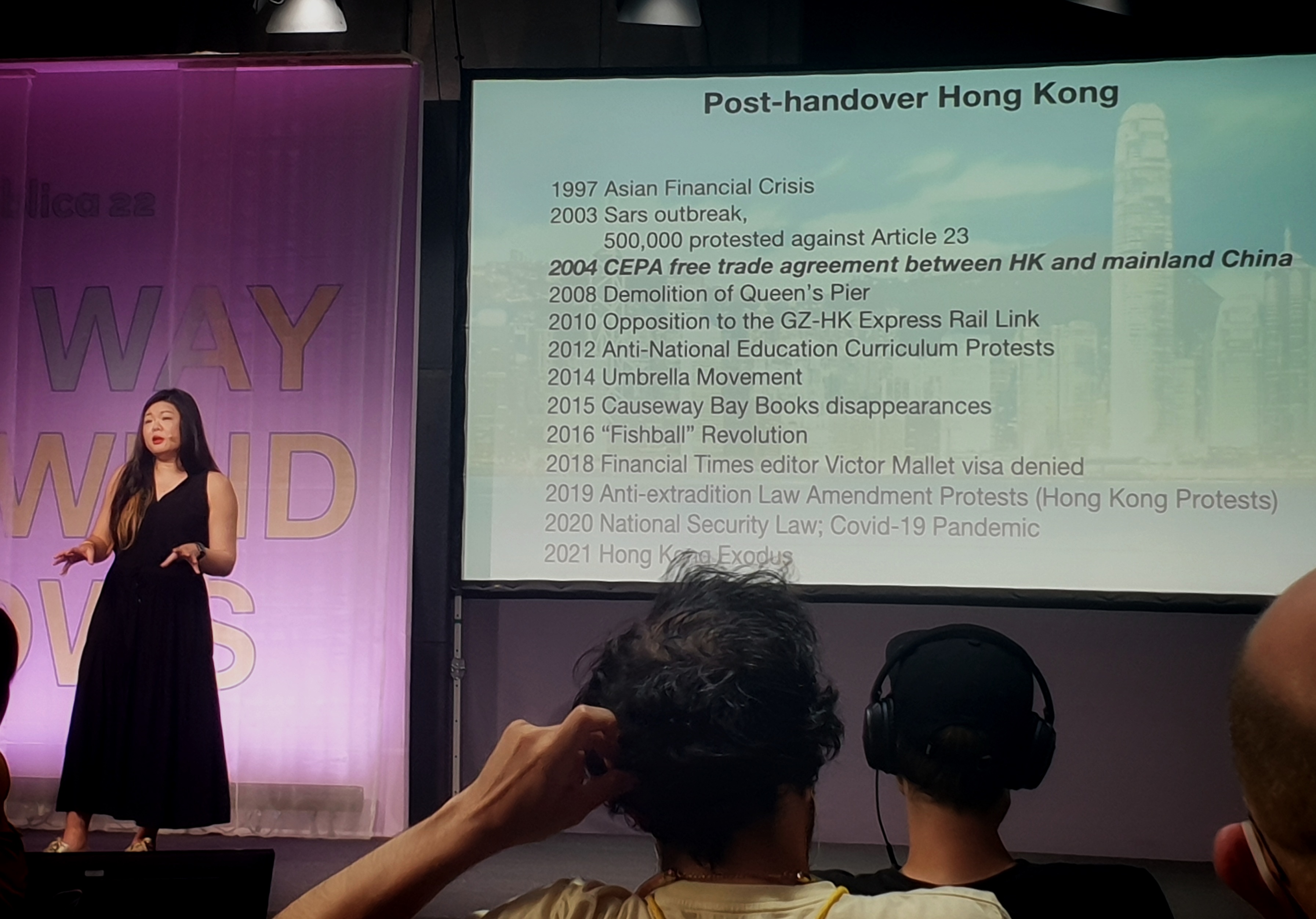 Vivienne Chow vor einer Timeline zu "Post-handover Hong Kong".
Die Daten: "1997 Asian Financial Crisis, 2003 Sars outbreak, 500.000 protested against Article 23, 2004 CEPA free trade agreement between HK and mainland China, 2008 Demolition of Queen's Pier, 2010 Opposition to the GZ-HK Express Rail Link, 2012 Anti-National Education Curriculum Protests, 2014 Umbrella Movement, 2015 Causeway Bay Books disappearances, 2016 "Fishball" Revolution, 2018 Financial Times editor Victor Mallet visa denied, 2019 Anti-extradition Law Amendment Protests (Hong Kong Protests), 2020 National Secutiry Law; Covid-19 Pandemic, 2021 Hong Kong Exodus"