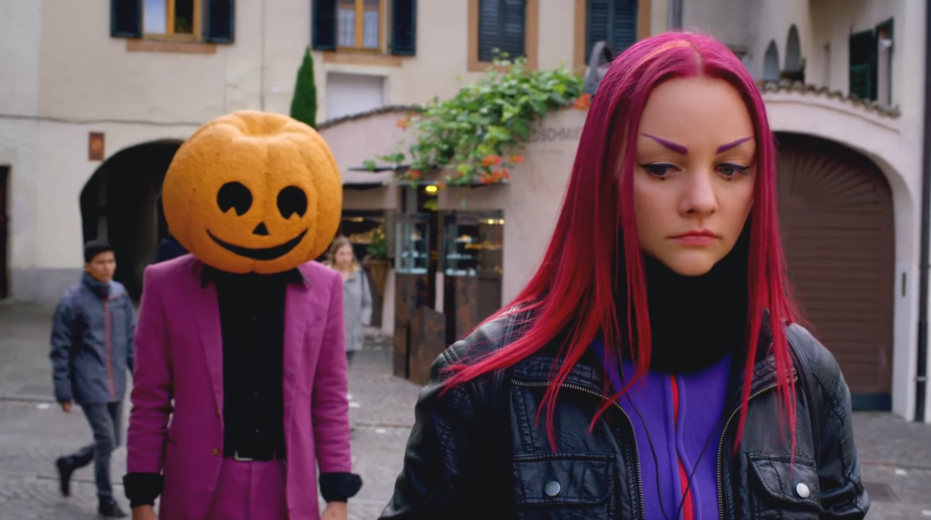 Ein weiblicher Teenager mit pinken Haaren, Lederjacke und traurigem Blick geht eine Straße entlang. Hinter ihr steht ein Mann mit Kürbiskopf und lila Anzug.
Im Hintergrund Gassen einer pittoresken italienischen Kleinstadt.