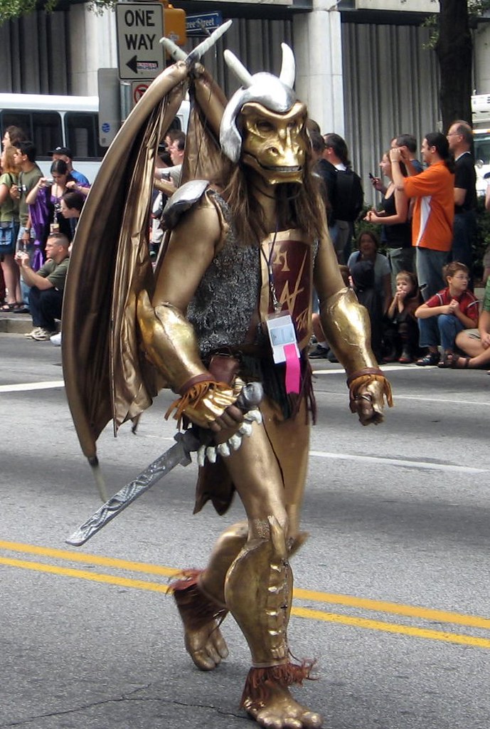 Ein Mensch im Drakonier-Kostüm mit braungoldenen Flügeln, Drachenmaske, silbernen Hörnern und goldenen Arm- und Beinschienen. Im Hintergrund viele Zuschauer.