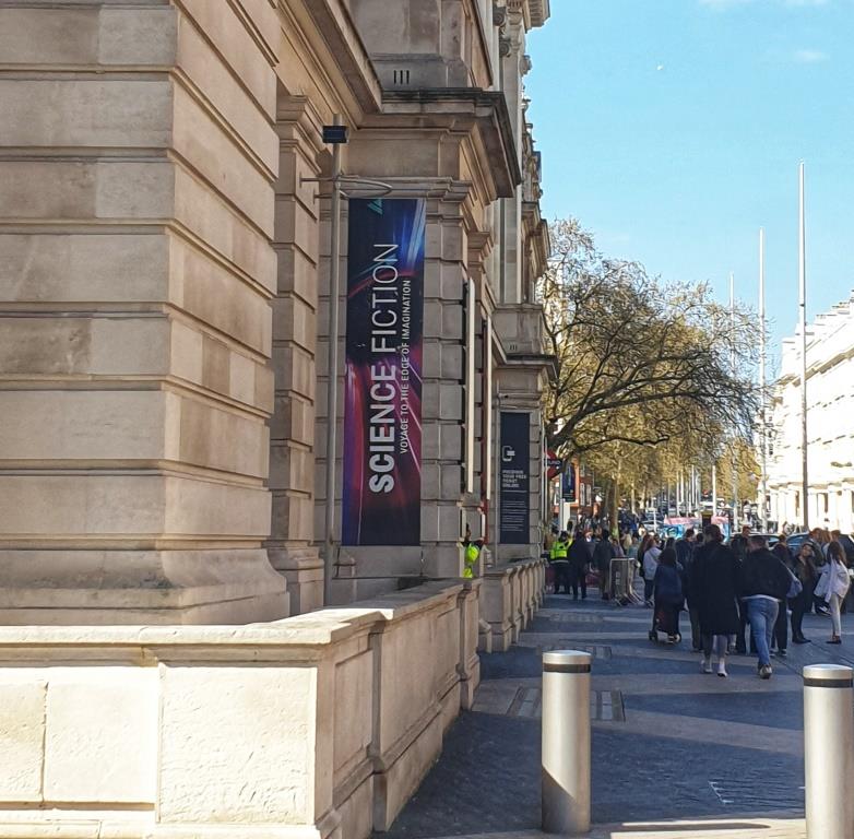 Fassade des Science Museum von außen. Ein klassizistischer Bau (?) bei Sonnenschein, viele Mensche stehen davor. Ein Plakat wirbt für die Ausstellung "Science Fiction"