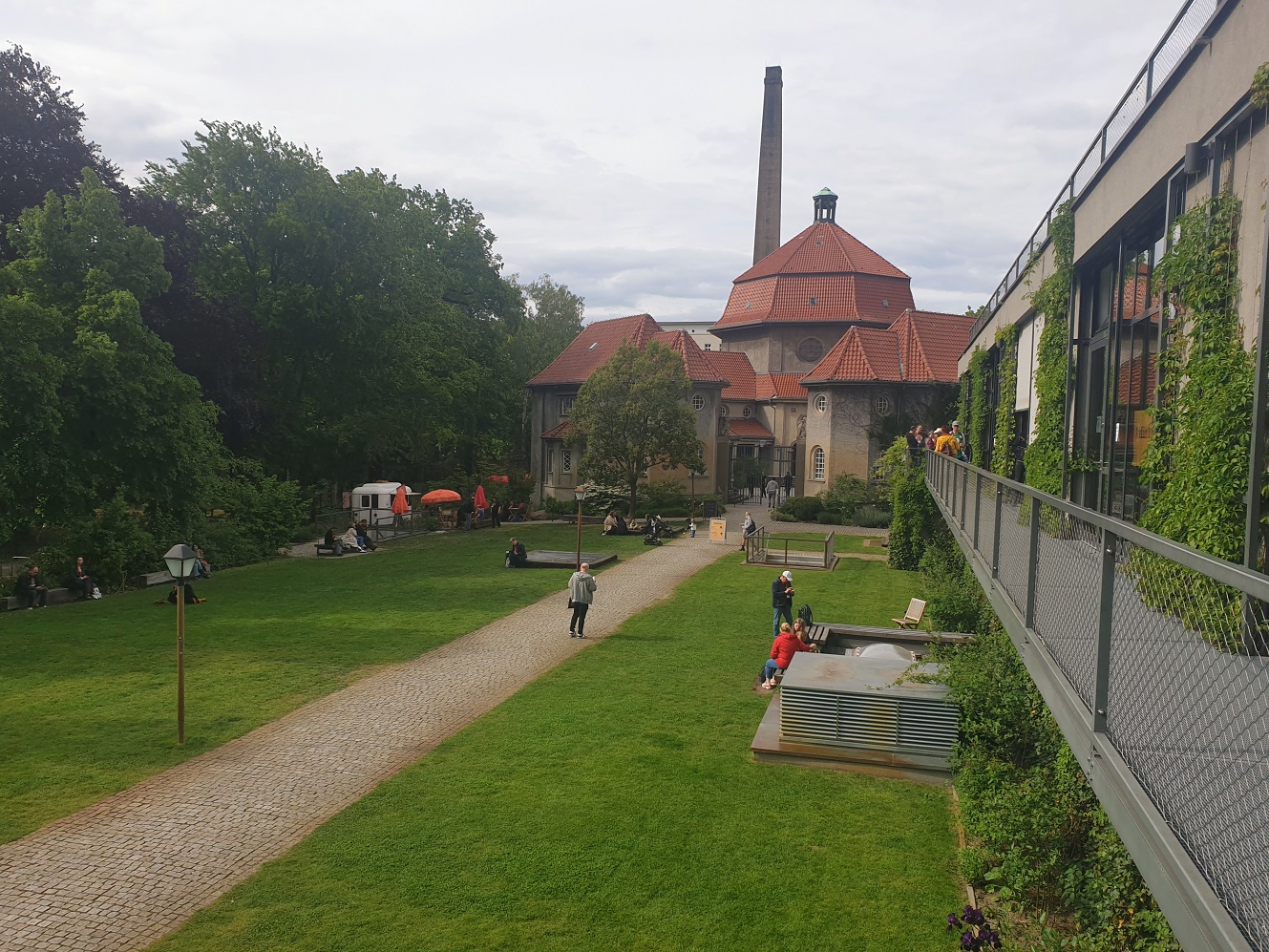 Ein weg zwischen grünen Wiesen, im Hintergrund ein altes Krematorium mit hohem Schornstein