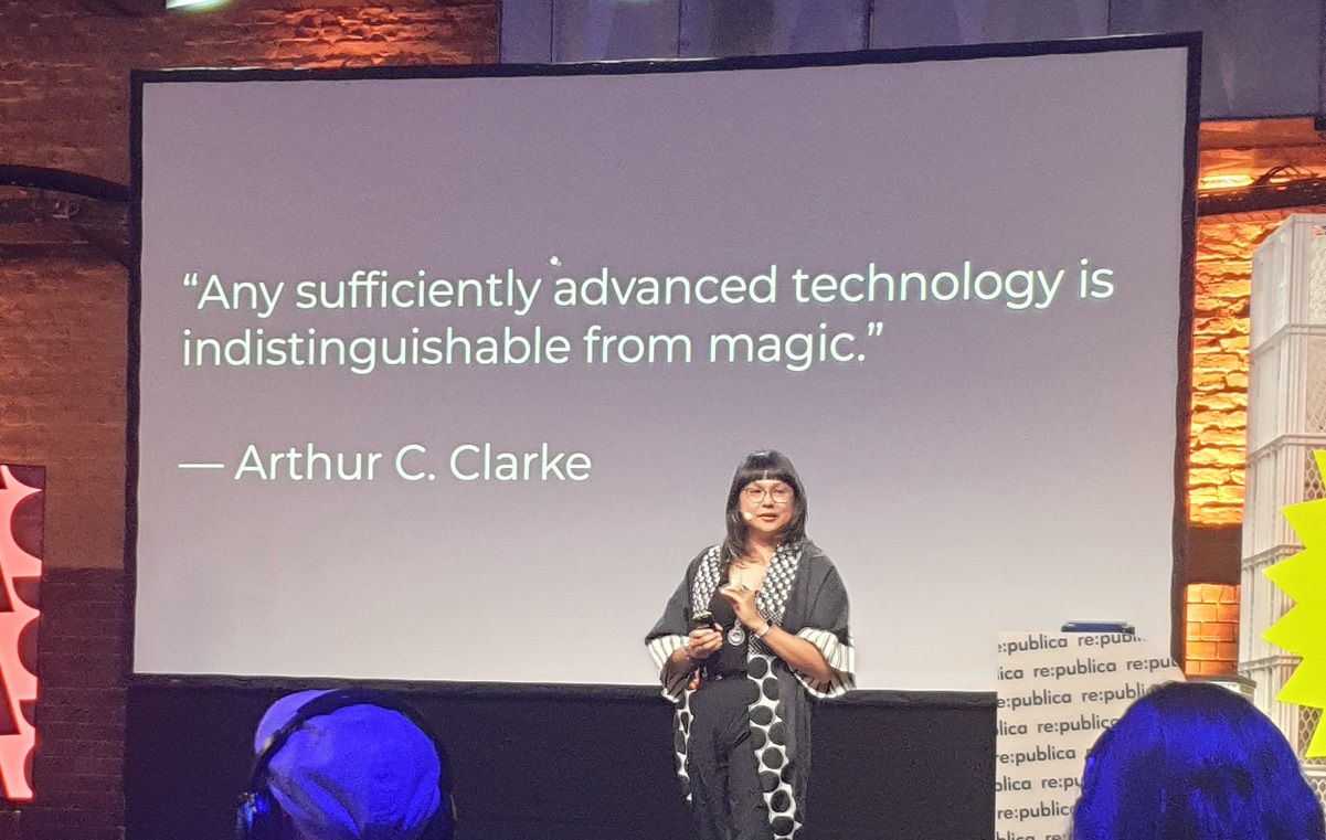 Braunhäutige Frau, AX Mina, mit schwarzen Haaren und Brillen referiert vor einem Bildschirm mit der Aufschrift "Any sufficiently advanced technology is indistinguishable from magic" - Arthur C. Clarke