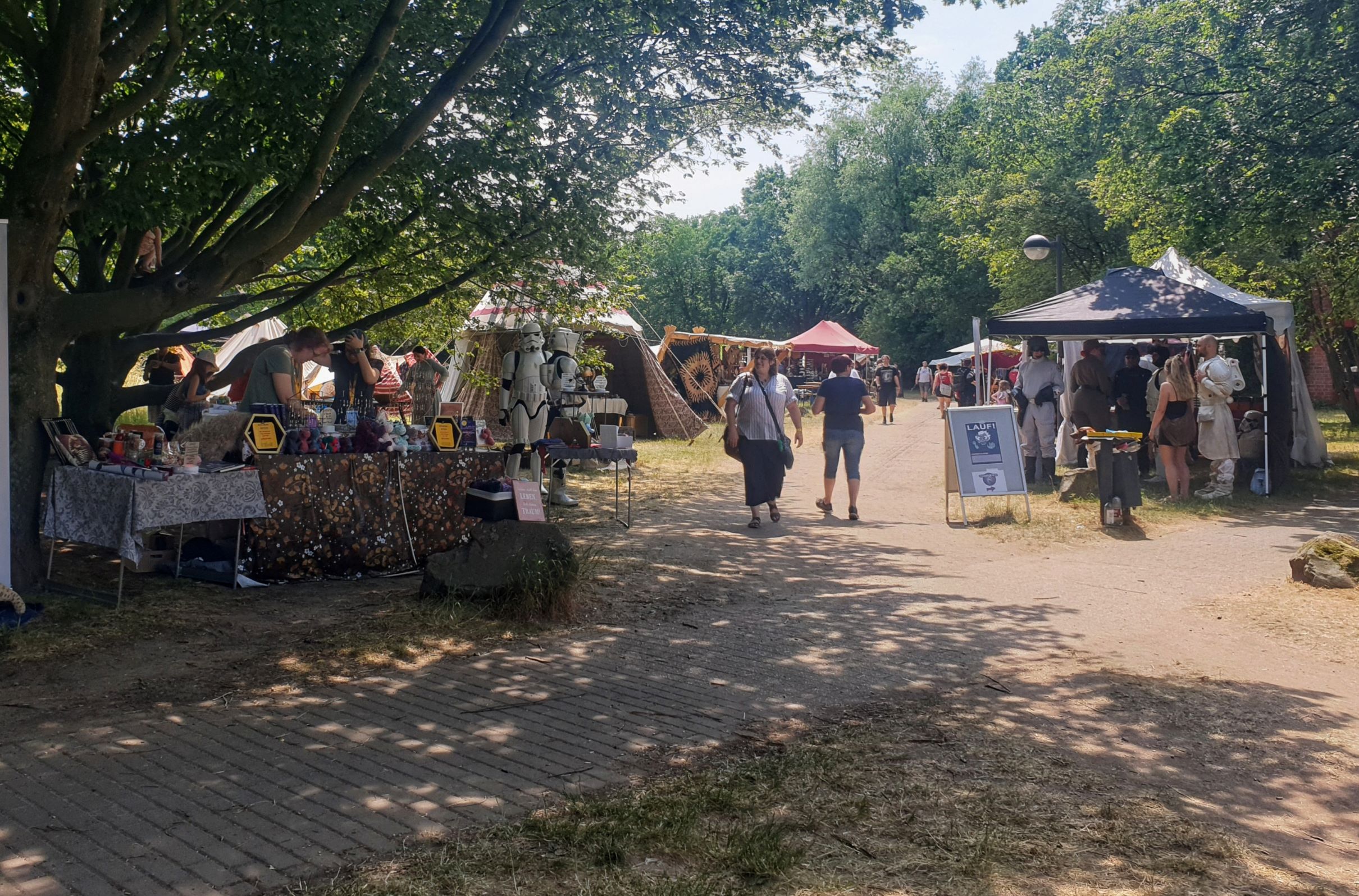 Marktstände auf einem Außengelände zwischen Bäumen an einem sonnigen Tag. In der Mitte steht ein Stormtropper.