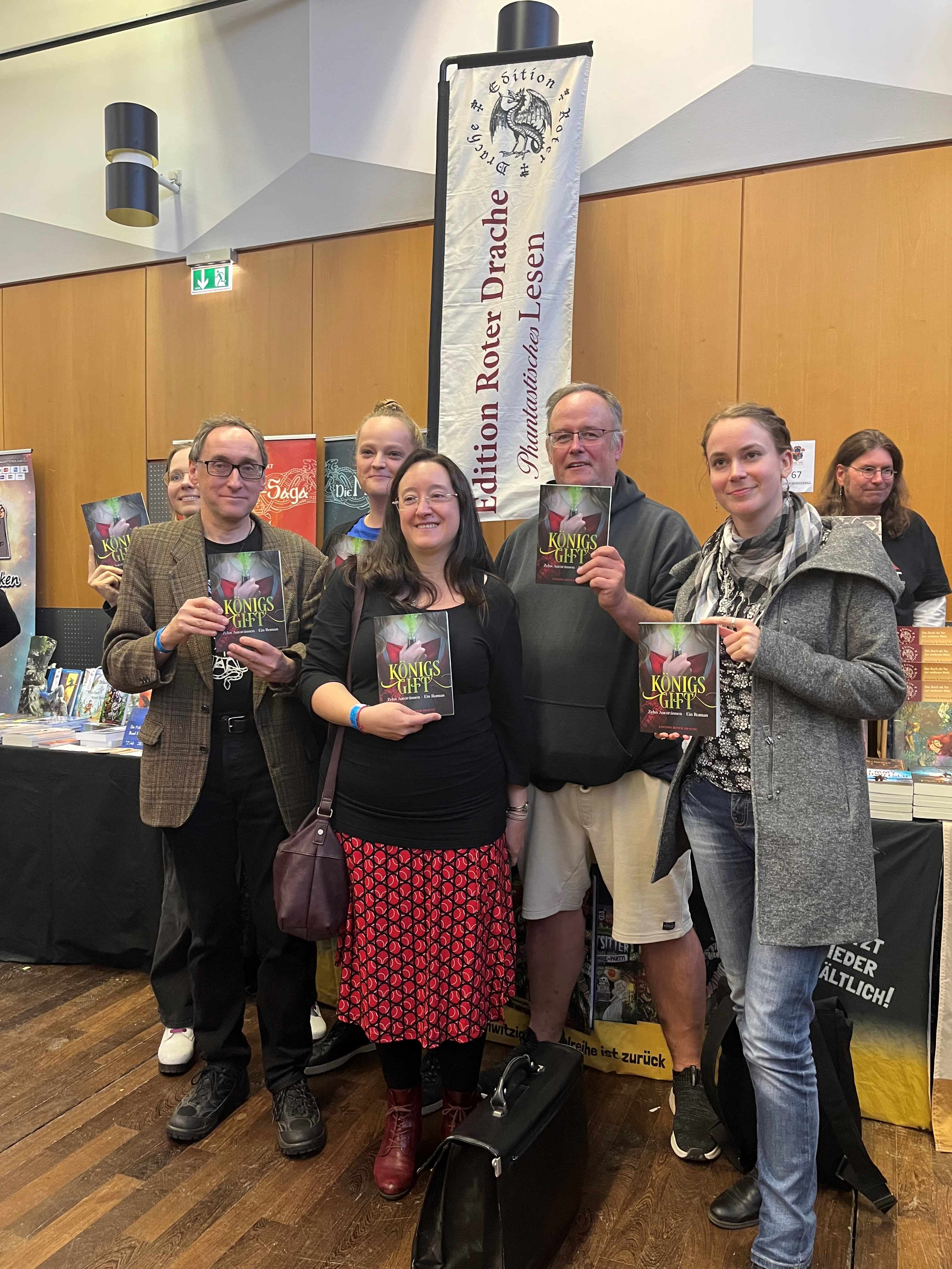 6 Autoren und Autorinnen halten vor einem Stand der "Edition Roter Drache" das Buch "Königsgift" hoch. In der Mitte Fabienne Siegmund, rechts von ihr Vincent Voss und Alessandra Ress. Links Theresa Hannig, Bernhard Stäber und etwas verdeckt Swantje Niemann