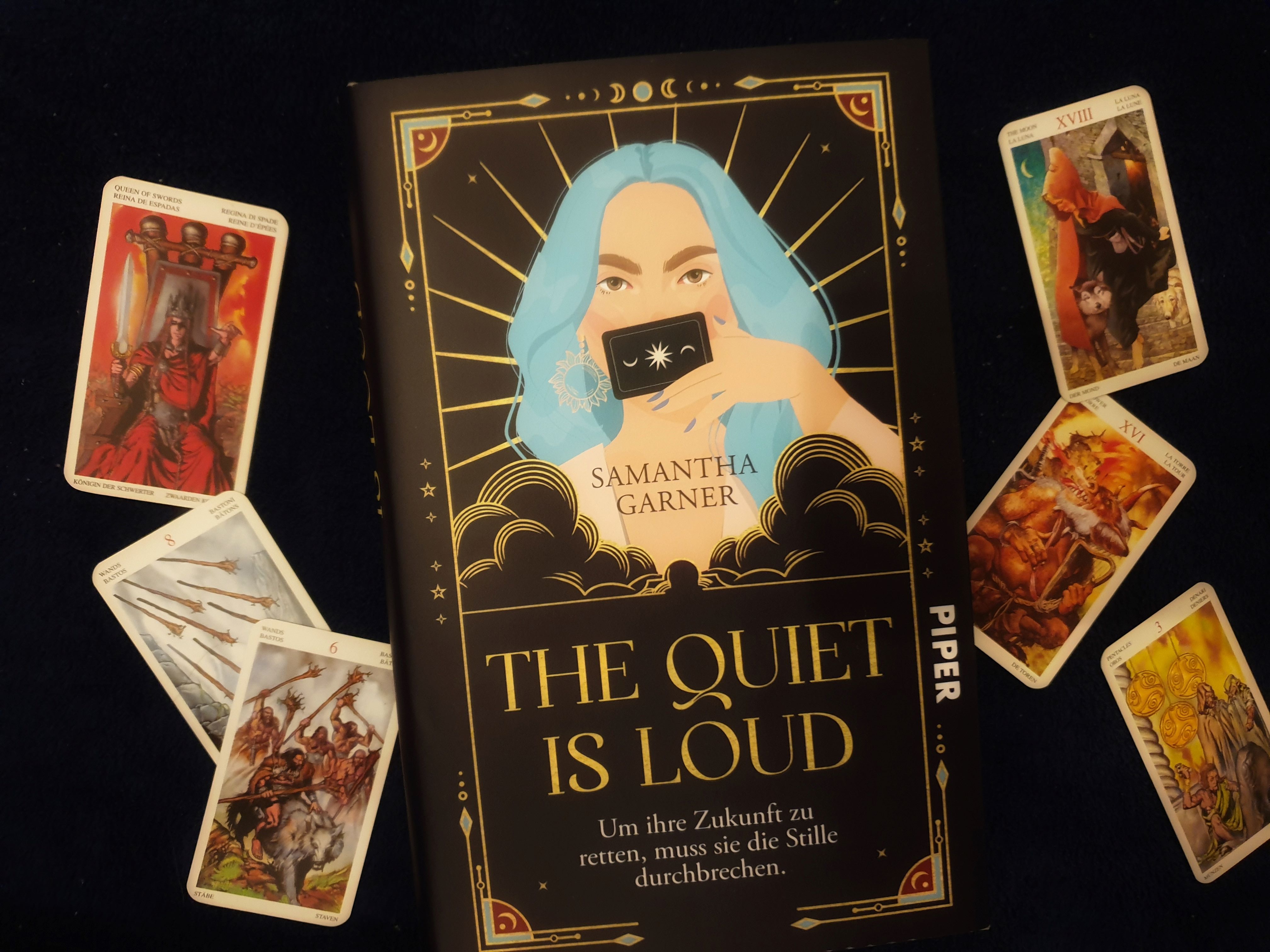 Buchcover von "The Quiet is Loud" zwoschen Tarotkarten. Das Cover zeigt eine blauhaarige Frau mit Tarotkarte.