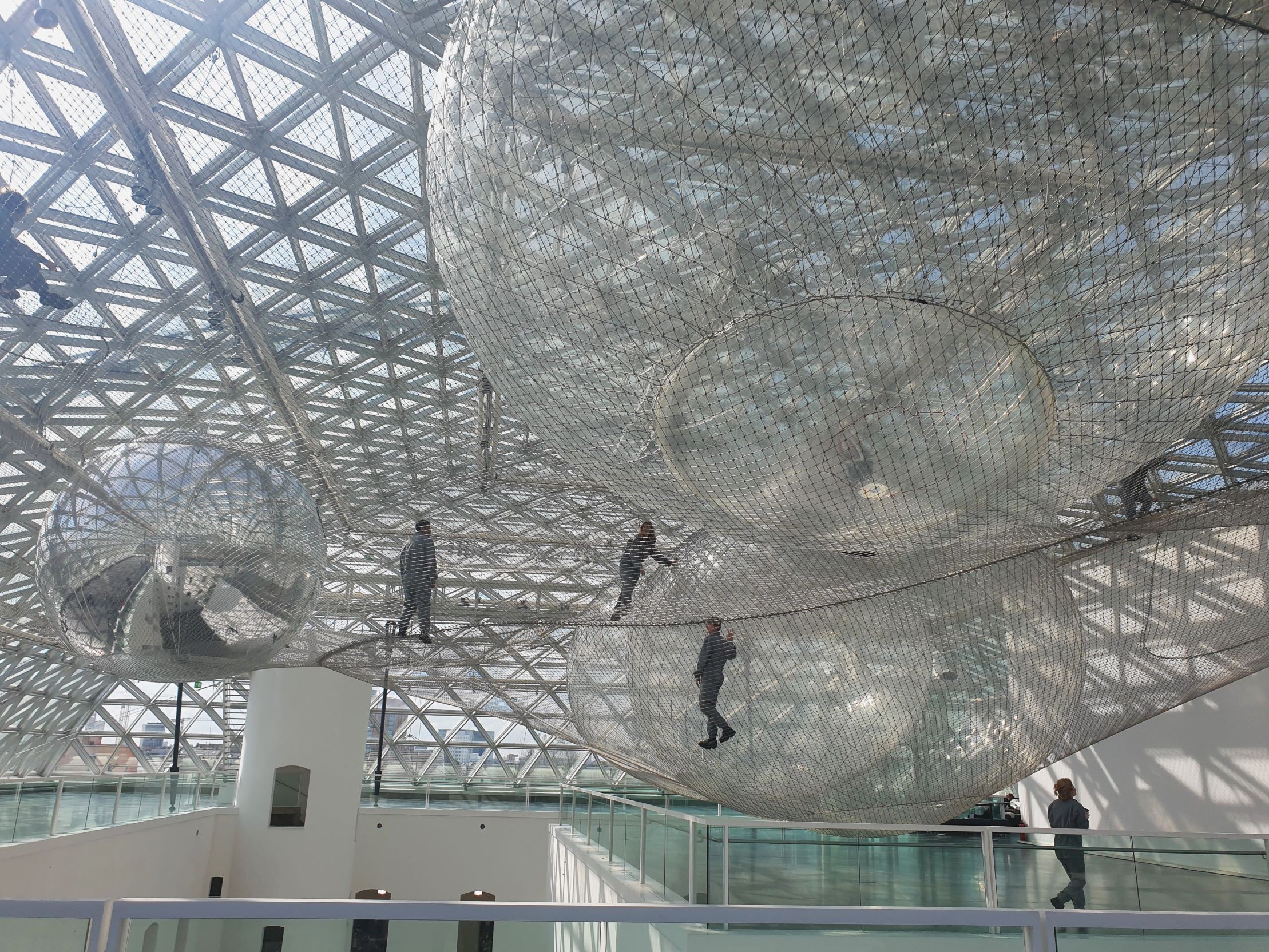 Kunstinstallation aus Netzen, die scheinbar Planeten unter dem sonnenbeschienen Dach bilden. Menschen in grauen Anzügen laufen auf den Netzen herum.