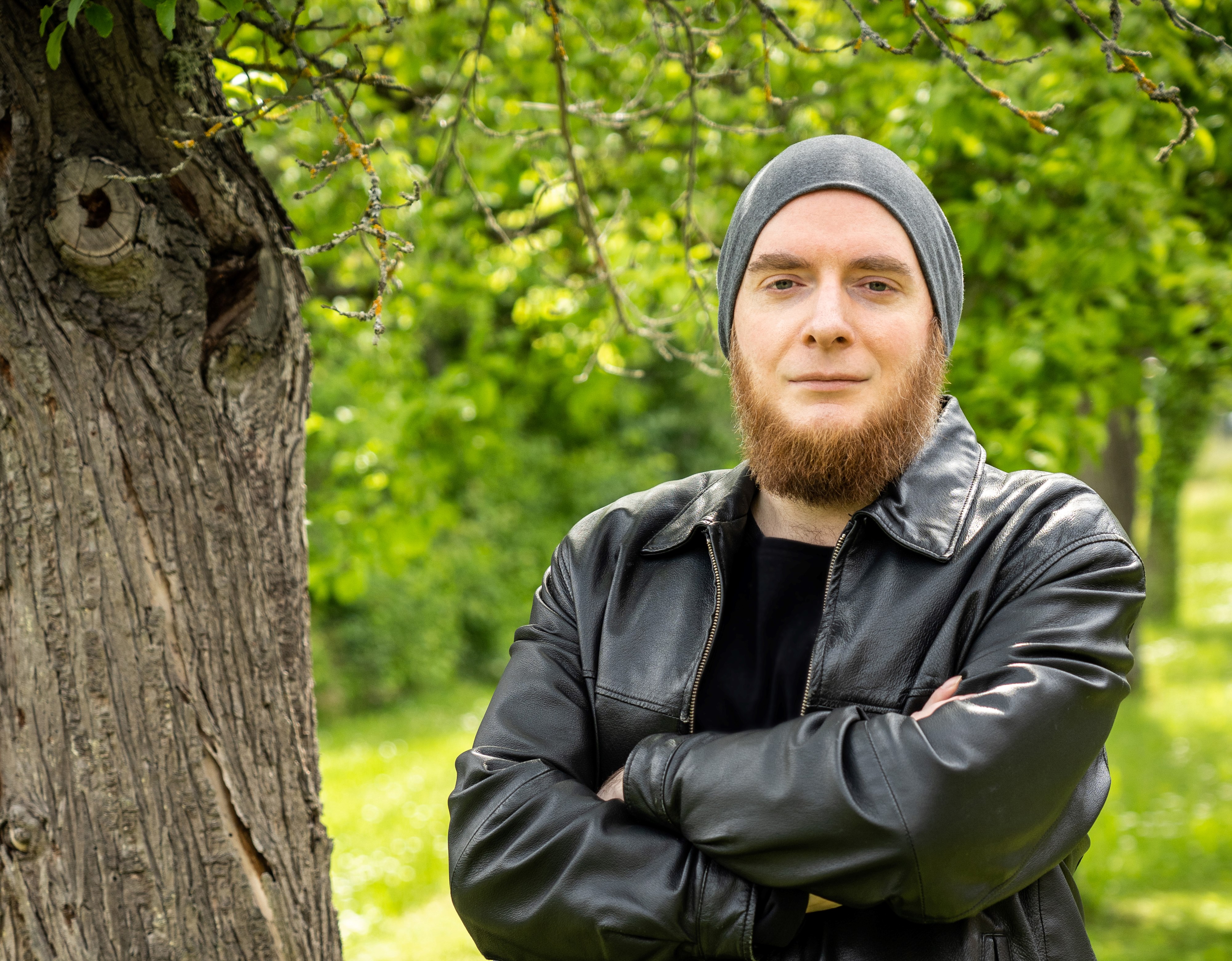 Mann mit rötlichem Backenbart, Mütze und Lederjacke steht mit verschränkten Armen neben einem Baum.