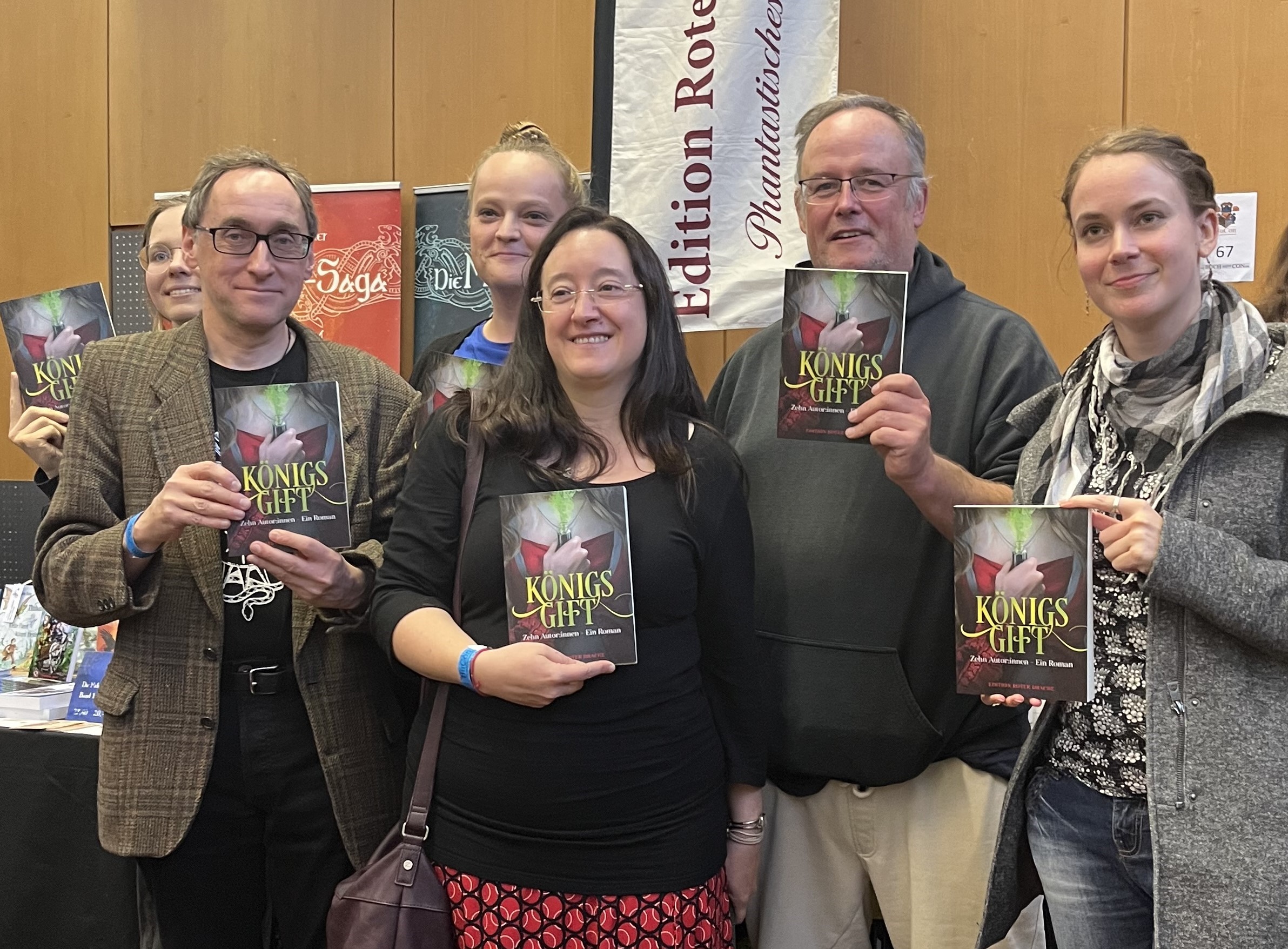 6 Autoren und Autorinnen halten vor einem Stand der "Edition Roter Drache" das Buch "Königsgift" hoch. In der Mitte Fabienne Siegmund, rechts von ihr Vincent Voss und Alessandra Ress. Links Theresa Hannig, Bernhard Stäber und etwas verdeckt Swantje Niemann