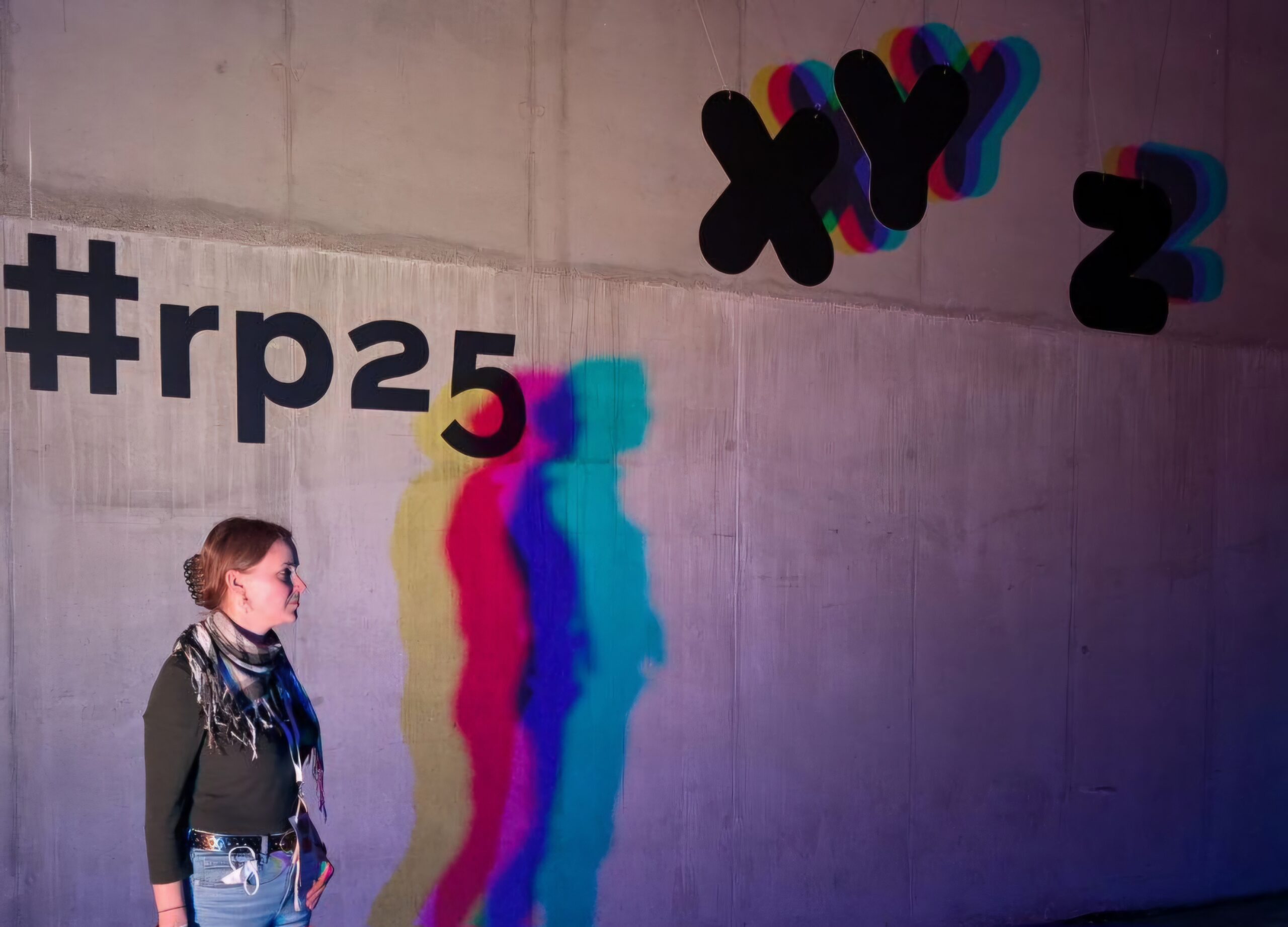 Alessandra Reß steht vor einer Wand mit der Aufschrift #rp25, drei bunte Schatten werden an die Wand geworfen