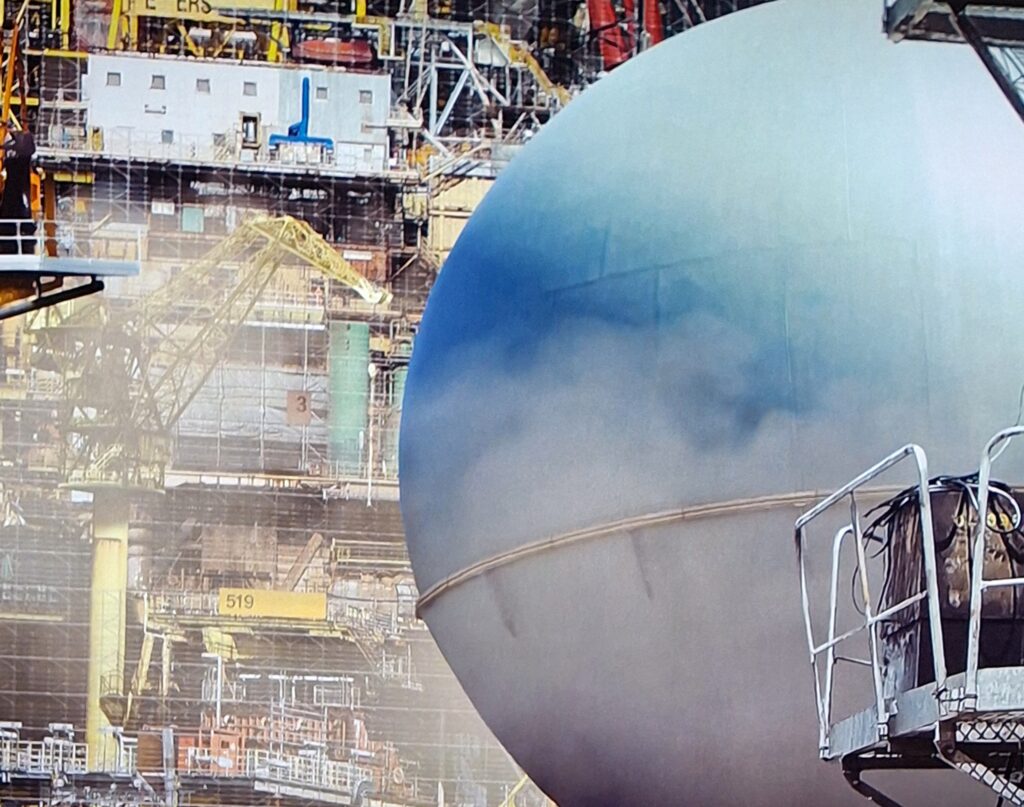 eine große blaue Kugel ähnlich einer Abrissbirne vor einer Art Baugerüst oder Containerschiff