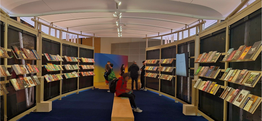 Blick in ein Holzgebäude, in dem viele Bücher ausgestellt sind. Eine Bank in der Mitte lädt zum Lesen ein.