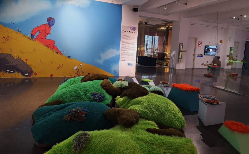 Einblick in die Ausstellung "Solarpunk" im Dortmunder U. Im Fokus ein großes Wandbild einer blauhäutigen Frau auf einem gelben Hügel und grüne "Hügel" als Sitzgelegenheiten