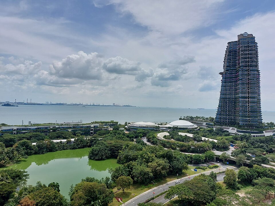 Blick auf eine grün bewachsene Stadt mit Wolkenkratzer auf der rechten Seite. Auf der linken Seite ein grüner See. Im Hintergrund das Meer oder ein breiter Fluss.
