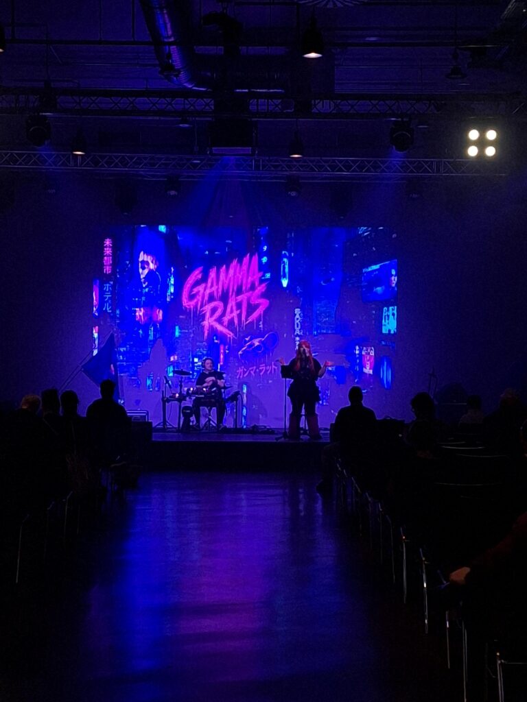 Foto der Gamma Rats. Sängerin und Drummer vor Cyberpunk-Ambiente, davor vage zu erkennen besetzte Stühle