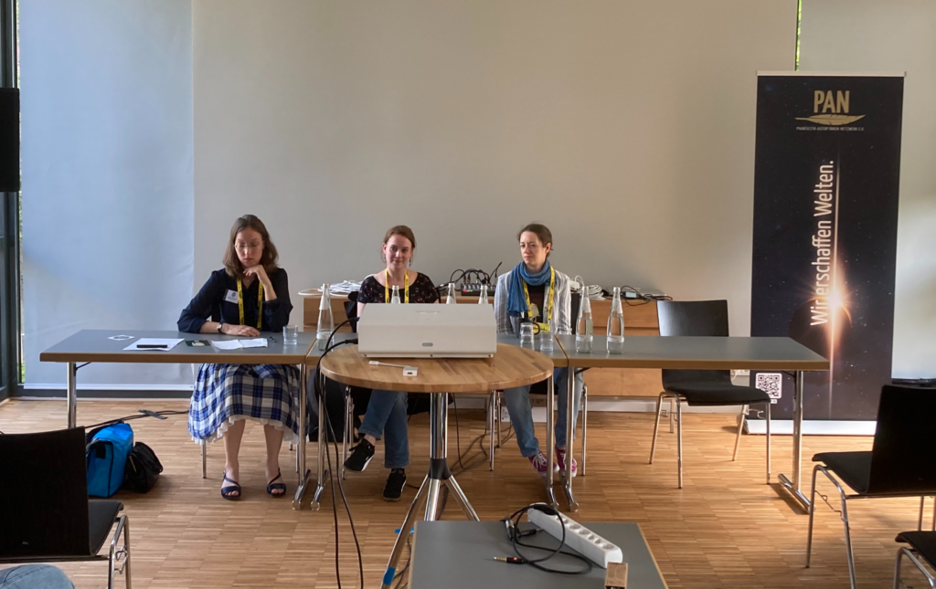 Hannah Brosch, Alessandra Reß und Christina Löw (drei blonde Frauen) sitzen an einem Tisch, vor sich Wassergläser und -flaschen. Ein Beamer steht in der Mitte, hinten ein PAN-Banner.
