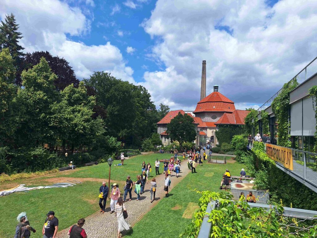 Menschen gehen einen Weg zwischen Wiesen entlang, an der Seite grün bewachsene Gebäudeteile und ein "Metropolcon"-Banner. Auf der Wiese sitzen und liegen ebenfalls Menschen. Im Hintergrund altes Krematorium mit rotem Dach