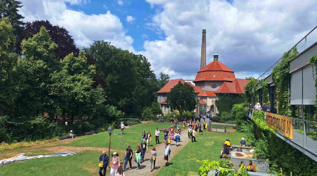 Menschen gehen einen Weg zwischen Wiesen entlang, an der Seite grün bewachsene Gebäudeteile und ein "Metropolcon"-Banner. Auf der Wiese sitzen und liegen ebenfalls Menschen. Im Hintergrund altes Krematorium mit rotem Dach