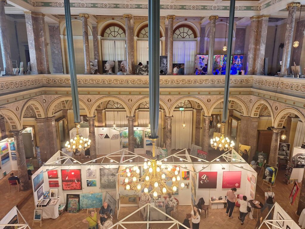 Blick von oben auf den mondänen Marmorsaal Bad Ems mit einer Ausstellung voller Gemälde. Drei Kronleuchter beleuchten die Ausstellung.