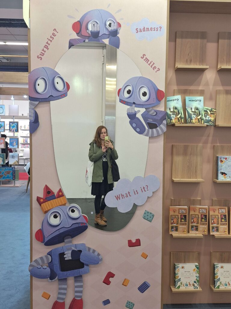 Spiegelselfie einer Frau vor einem Spiegel mit vier Robotern und Sprechblasen: "Surprise?", "Sadness?", "Smile?", "What is it?"